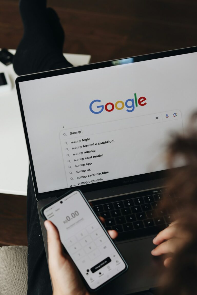 A person is using a laptop to search for SumUp on Google while holding a white coffee mug. The Google search page is displayed on the screen, and the user is preparing to click Google Search.